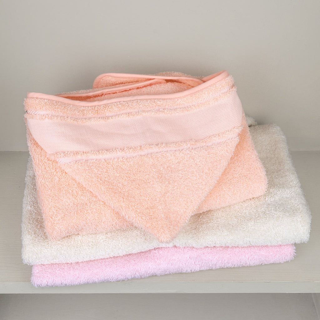Cape de bain bébé à capuche en éponge coton longues fibres, fabrication portugaise, coloris saumon, écru et rose, rangée sur une étagère