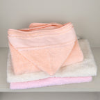 Cape de bain bébé à capuche en éponge coton longues fibres, fabrication portugaise, coloris saumon, écru et rose, rangée sur une étagère