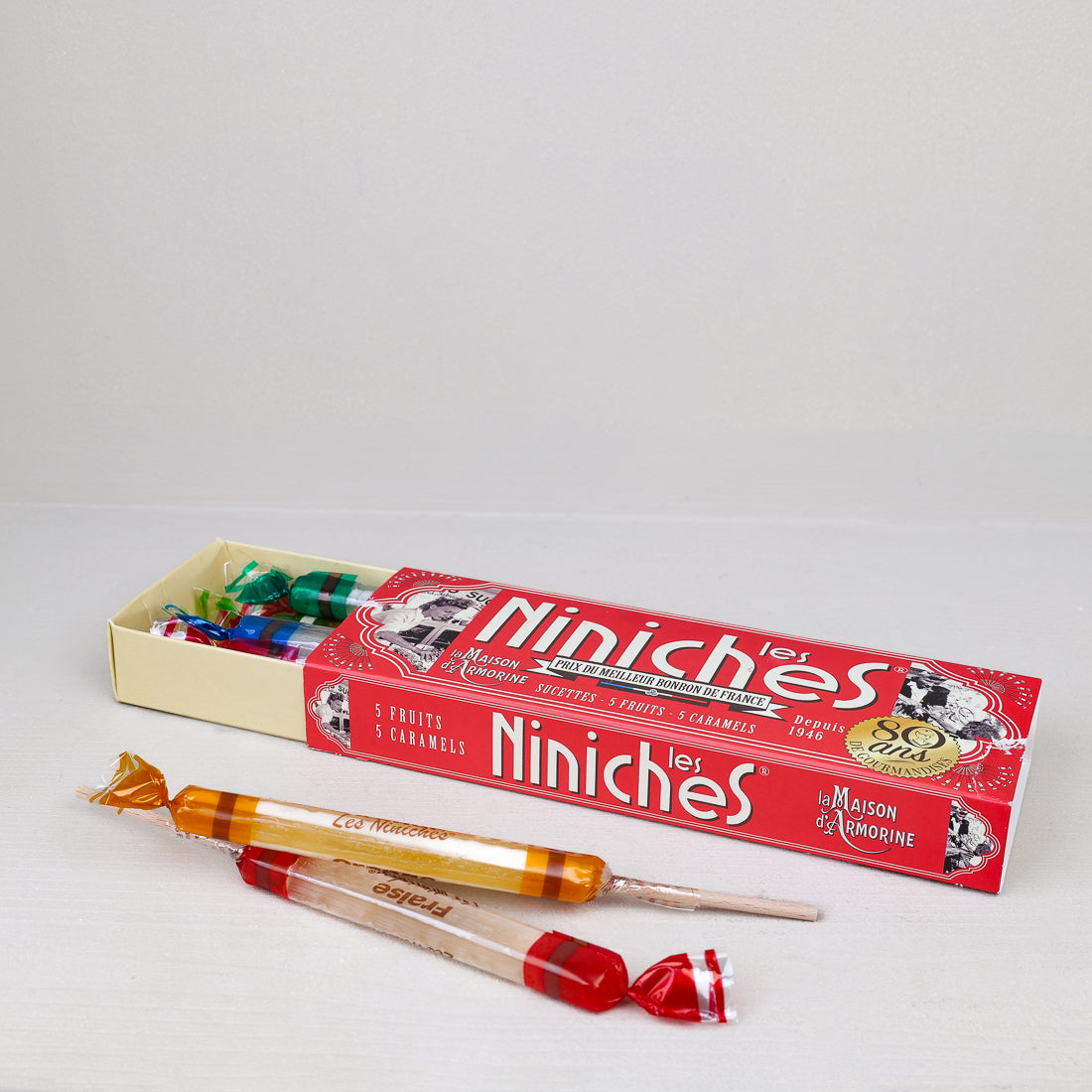 Étui ouvert Les Niniches Maison Armorine 160g avec sucettes caramel et fruits posées devant, confiserie bretonne anniversaire 80 ans